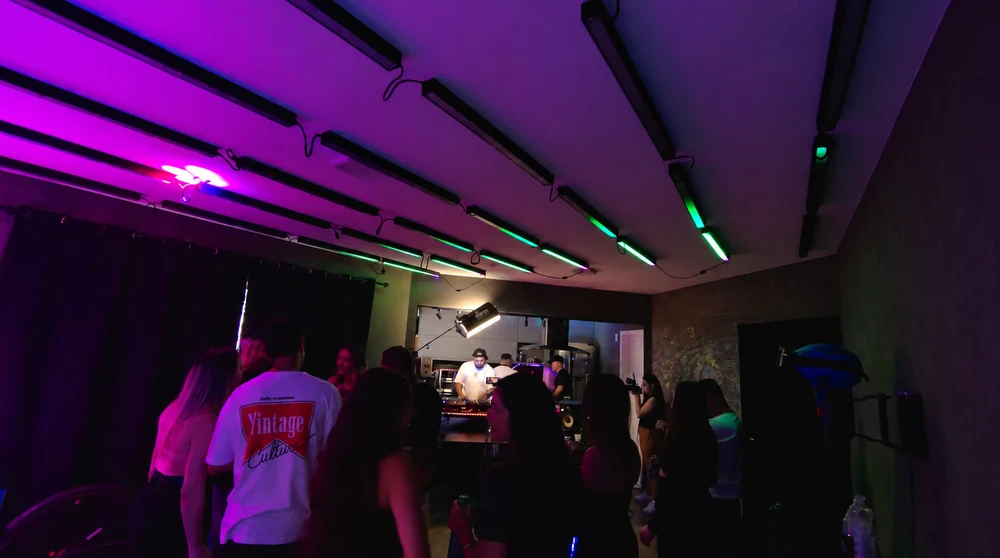 Convidados no apartamento sob luzes magenta e verde, com Haquino na cozinha ao fundo